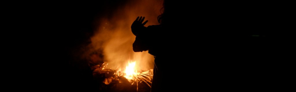 Drew Mirandus, in front of a campfire, raising hand to feel the heat.