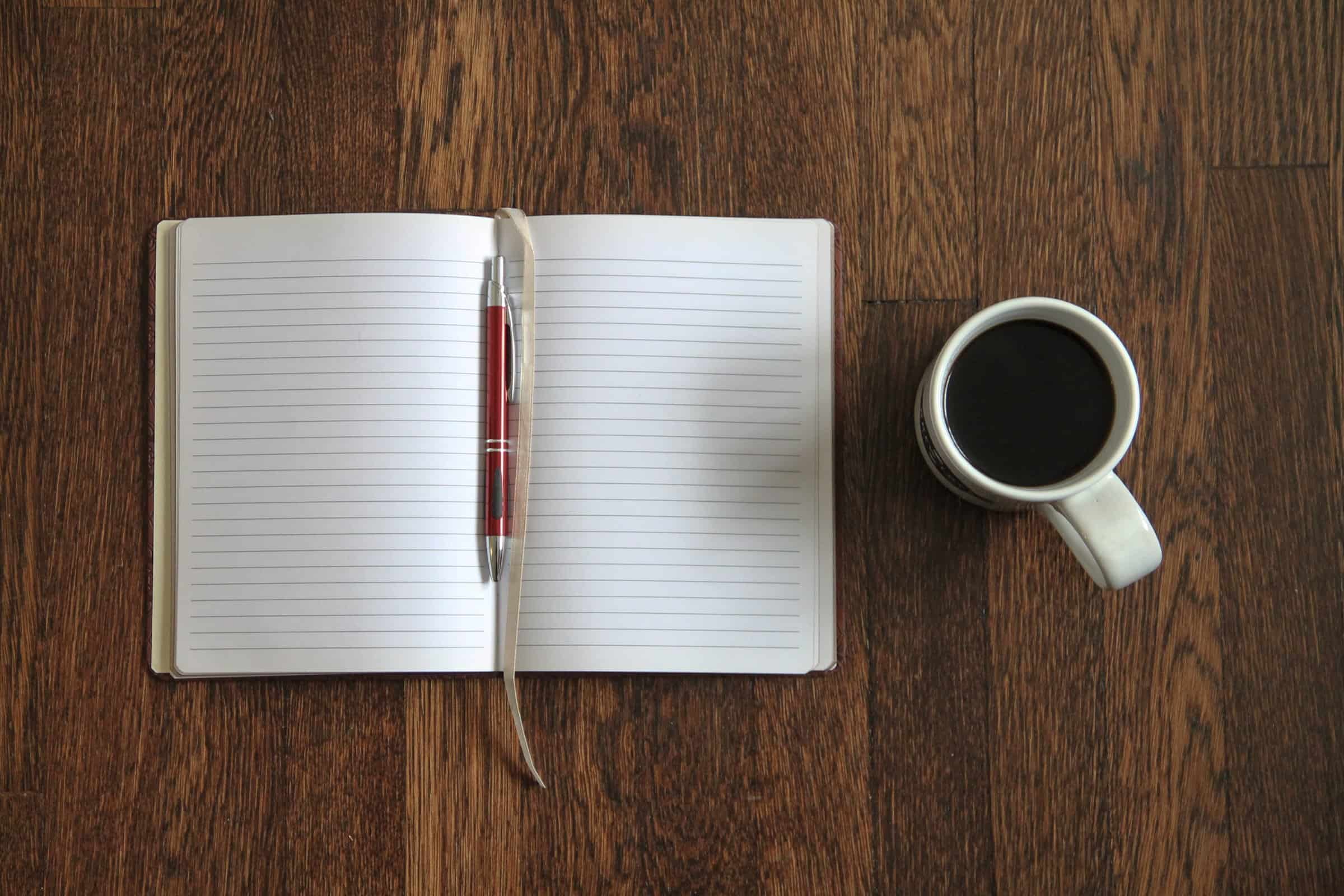 Open notebook with a red pen and a cup of black coffee on a wooden desk. Stock photo from Good Stock Photos.