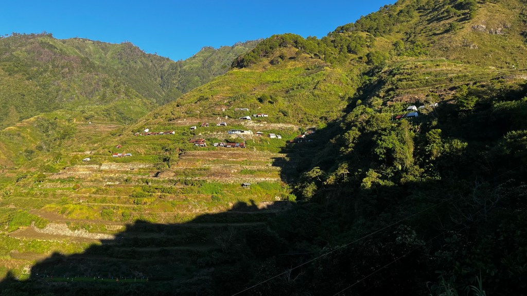 A distant view of Buscalan Village nestled in the Cordillera mountains, surrounded by lush greenery and misty peaks, capturing the natural beauty and cultural essence of the region.