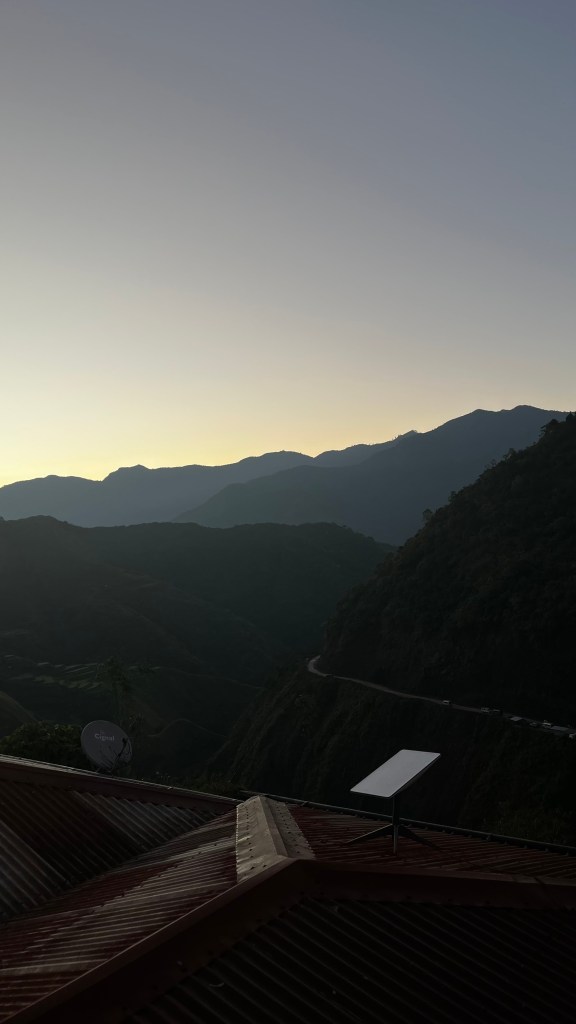 A serene view of the mountains surrounding Buscalan Village at the break of dawn, with the sky transitioning from dark to light. The rooftops and satellite dishes are visible in the foreground.