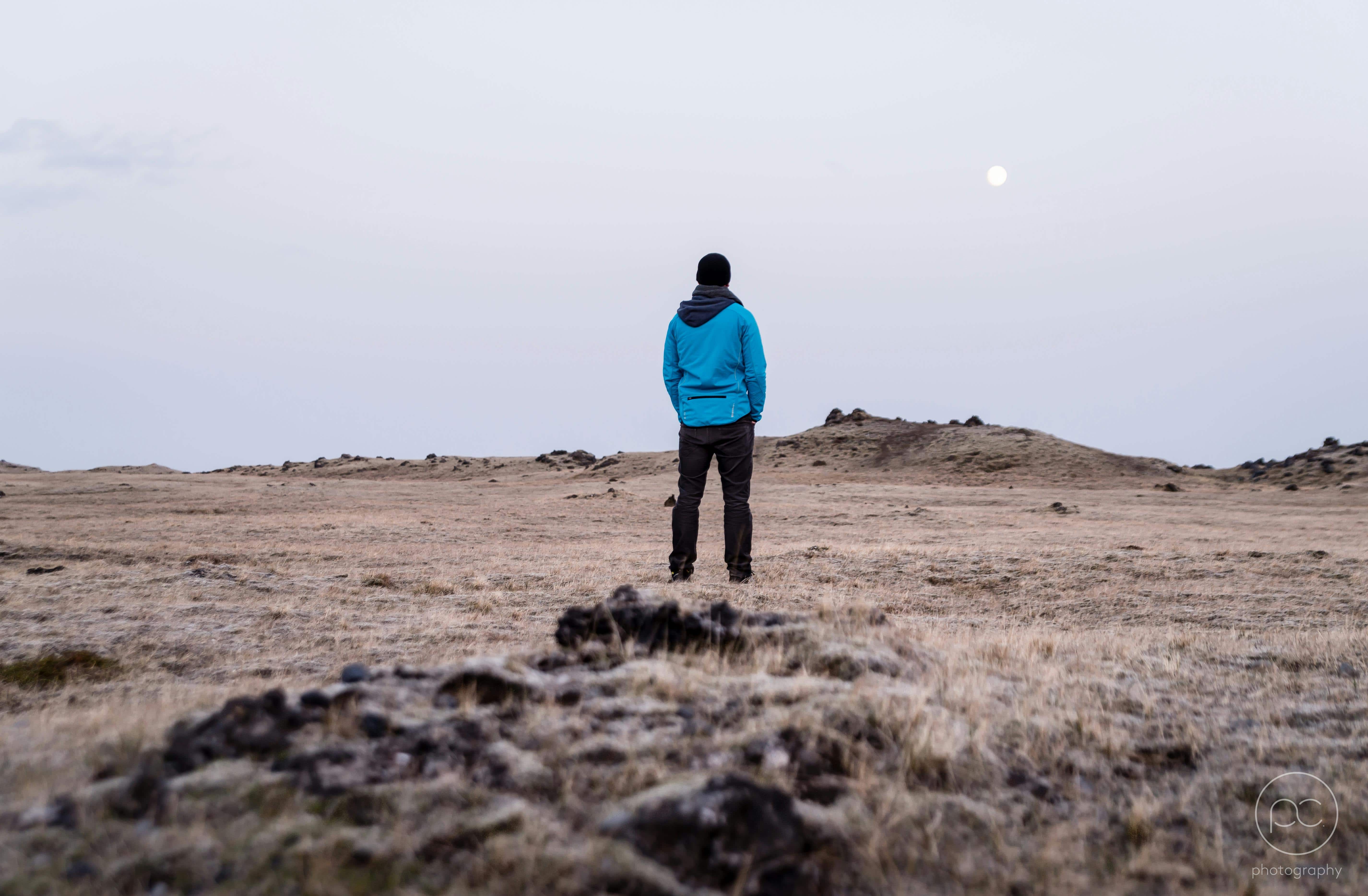 A person in a blue jacket stands alone in a barren field under a pale sky with the moon overhead, symbolizing solitude, reflection, and the emotional clarity of solo travel.