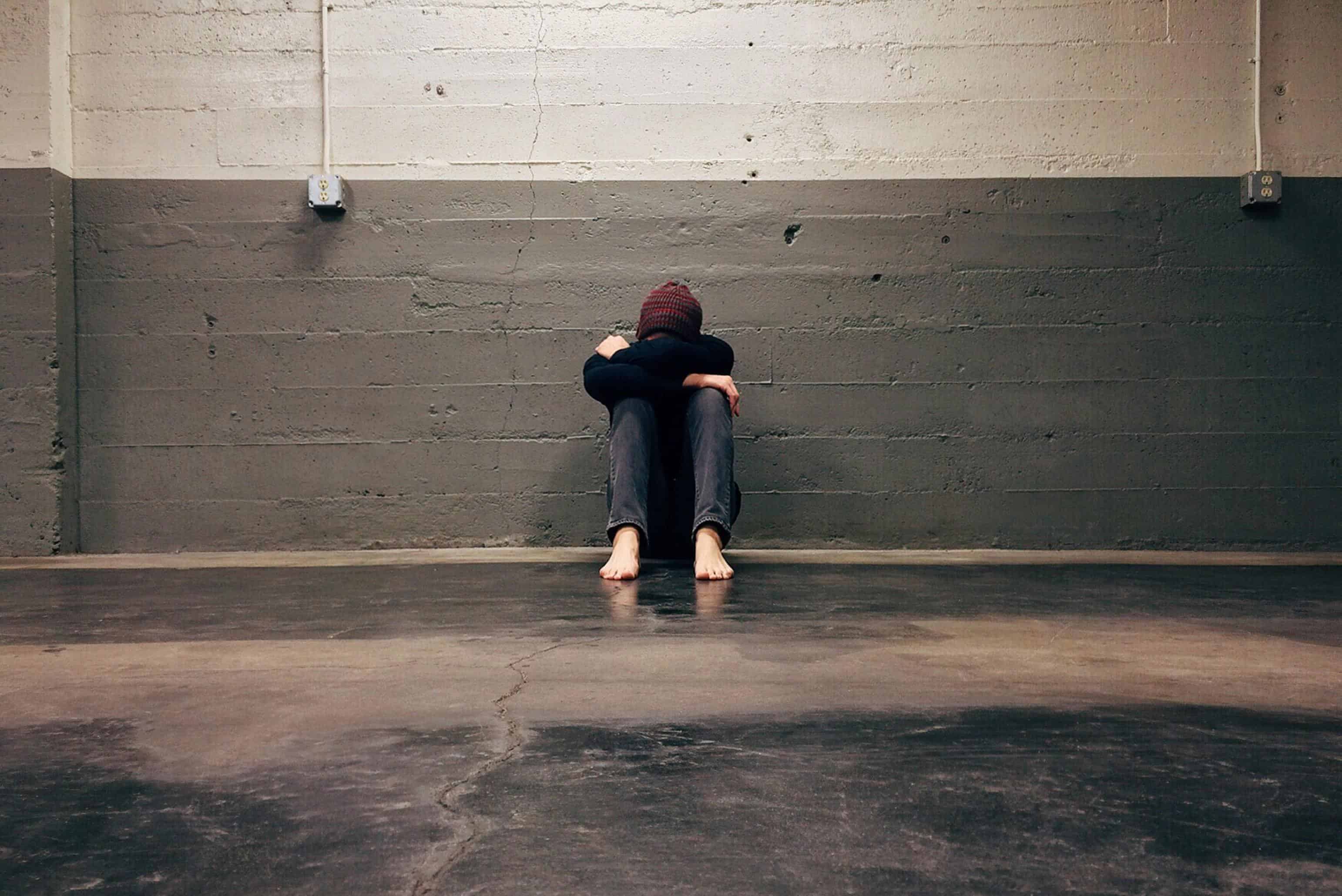Person sitting barefoot against a concrete wall, curled into themselves in a posture of emotional exhaustion and spiritual despair.