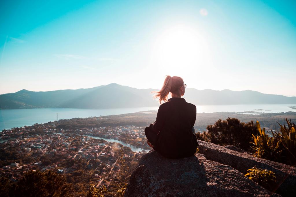 Person sitting alone on a mountain cliff, overlooking a coastal town and calm sea during sunrise, reflecting in solitude