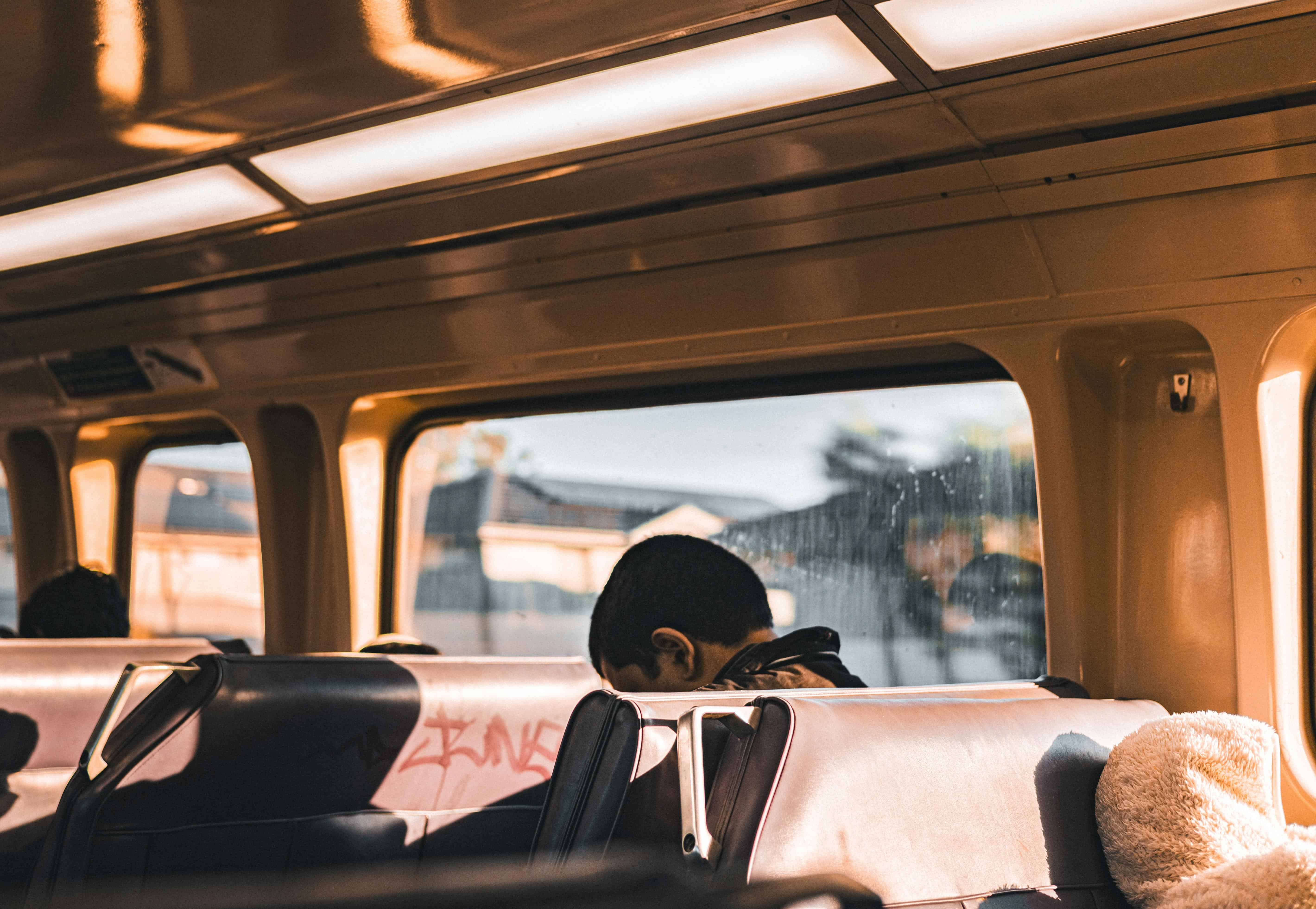 Person sitting alone on a train with head lowered, sunlight streaming in through the windows