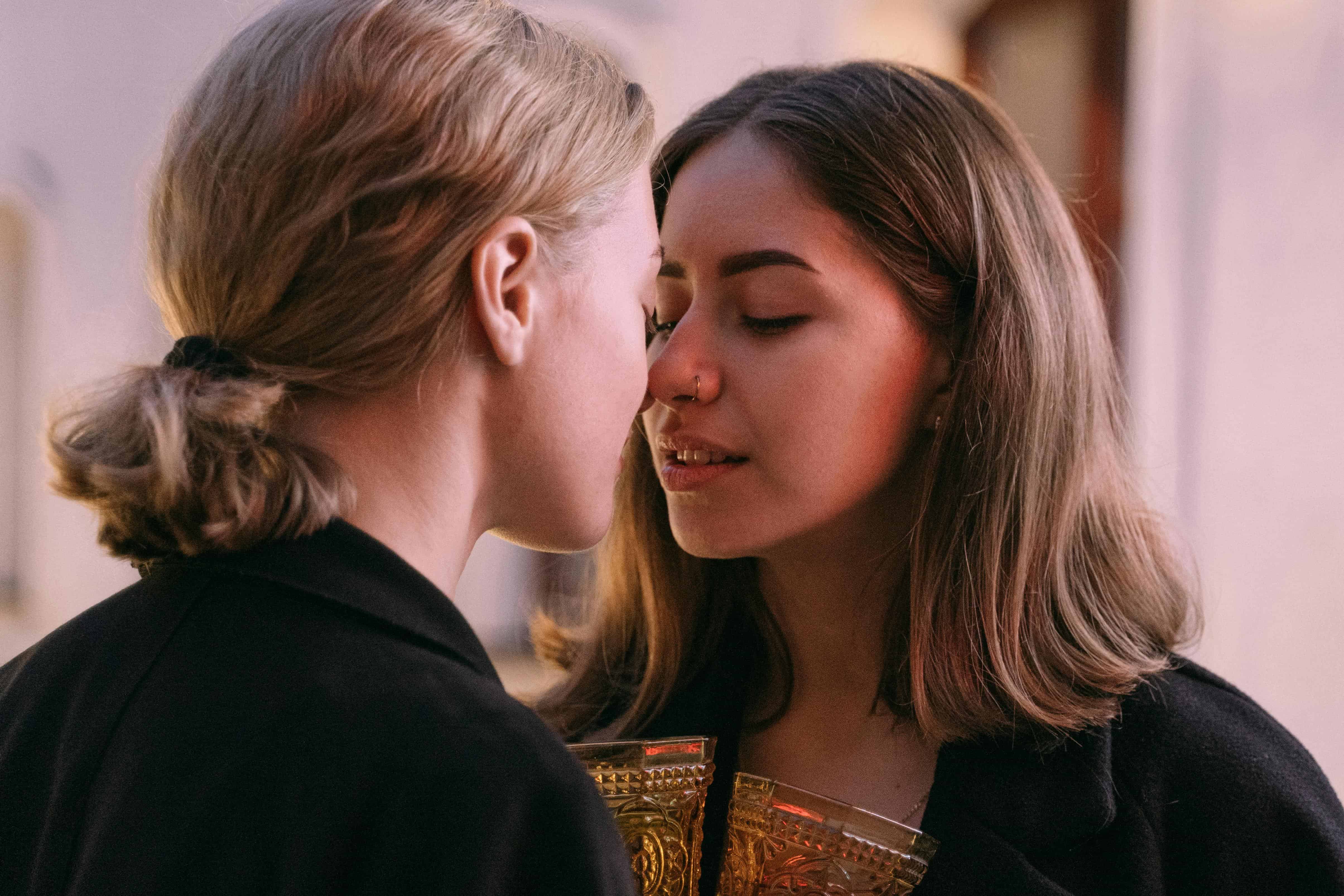 Two women leaning in closely, eyes closed, holding golden glasses and sharing an intimate, vulnerable moment
