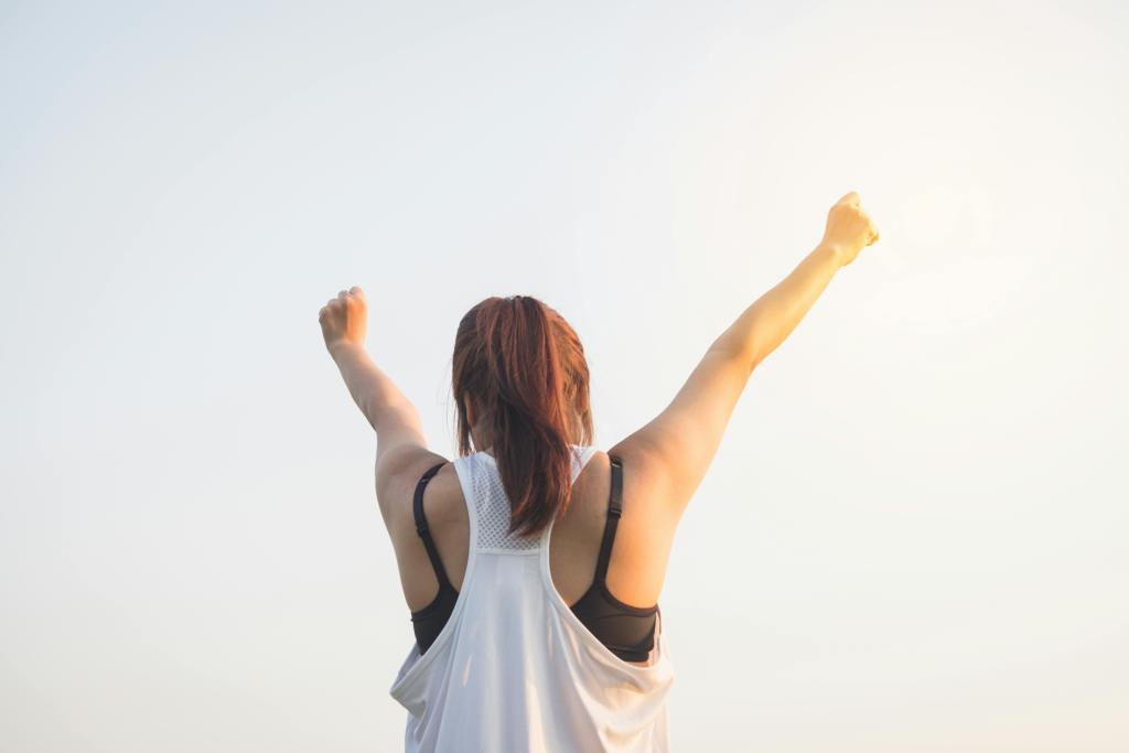 Back view of a woman with arms raised in victory under a clear sky, symbolizing self-trust and personal strength