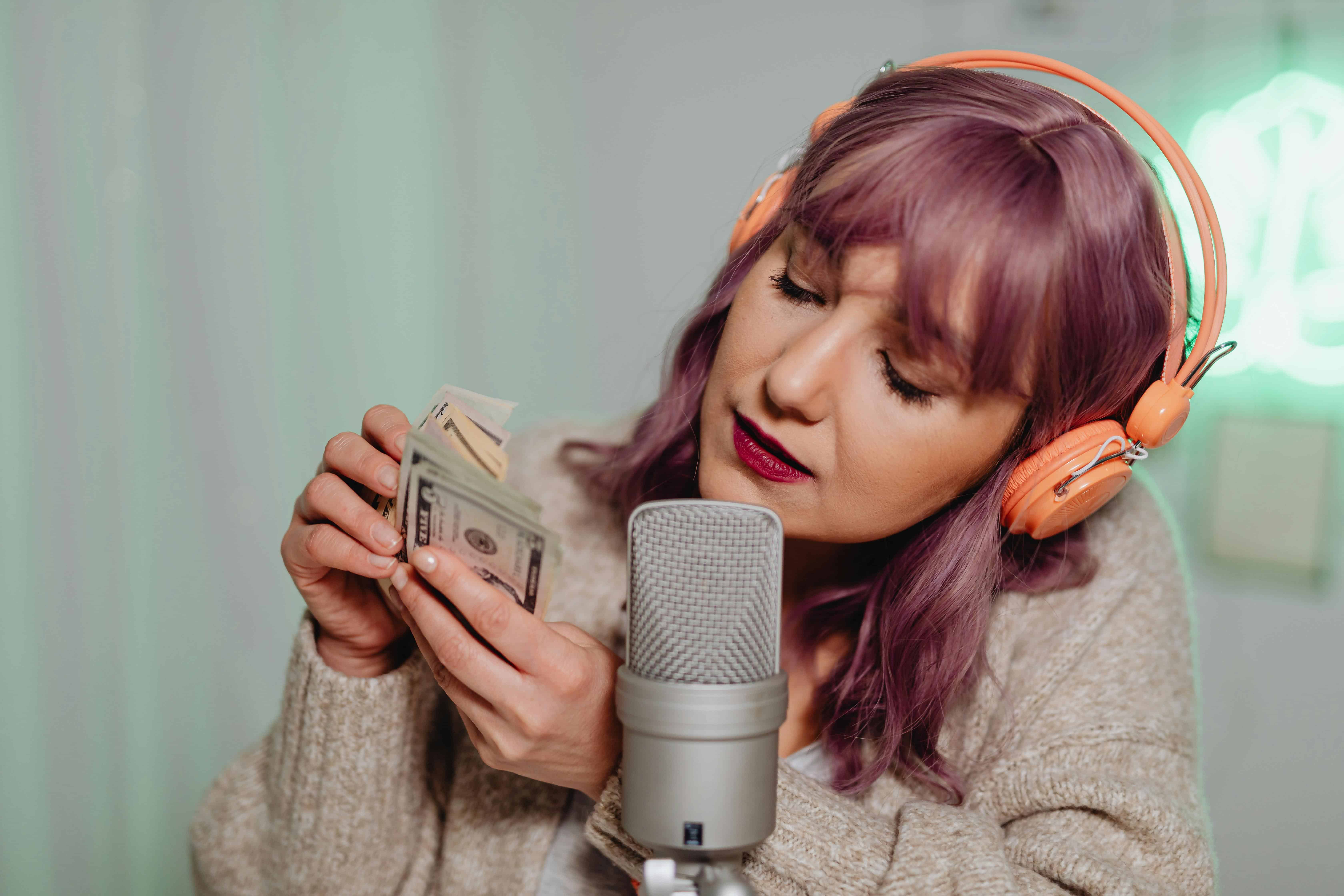 Woman with purple hair wearing orange headphones and recording ASMR sounds by counting money into a microphone.