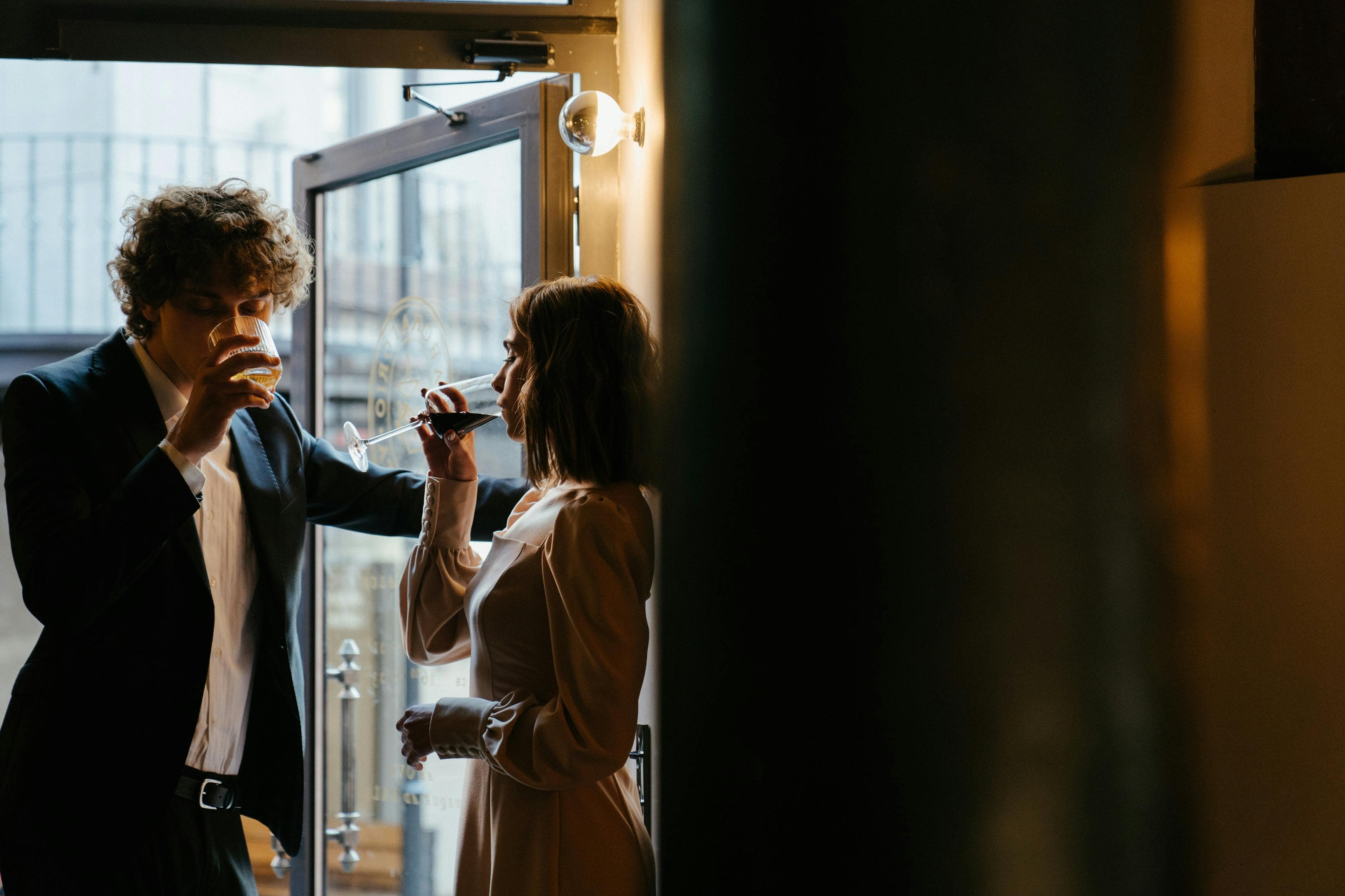 A man in a suit and a woman in a cream dress share drinks by a glass door, capturing the essence of an organic meeting that feels like a modern meet cute.