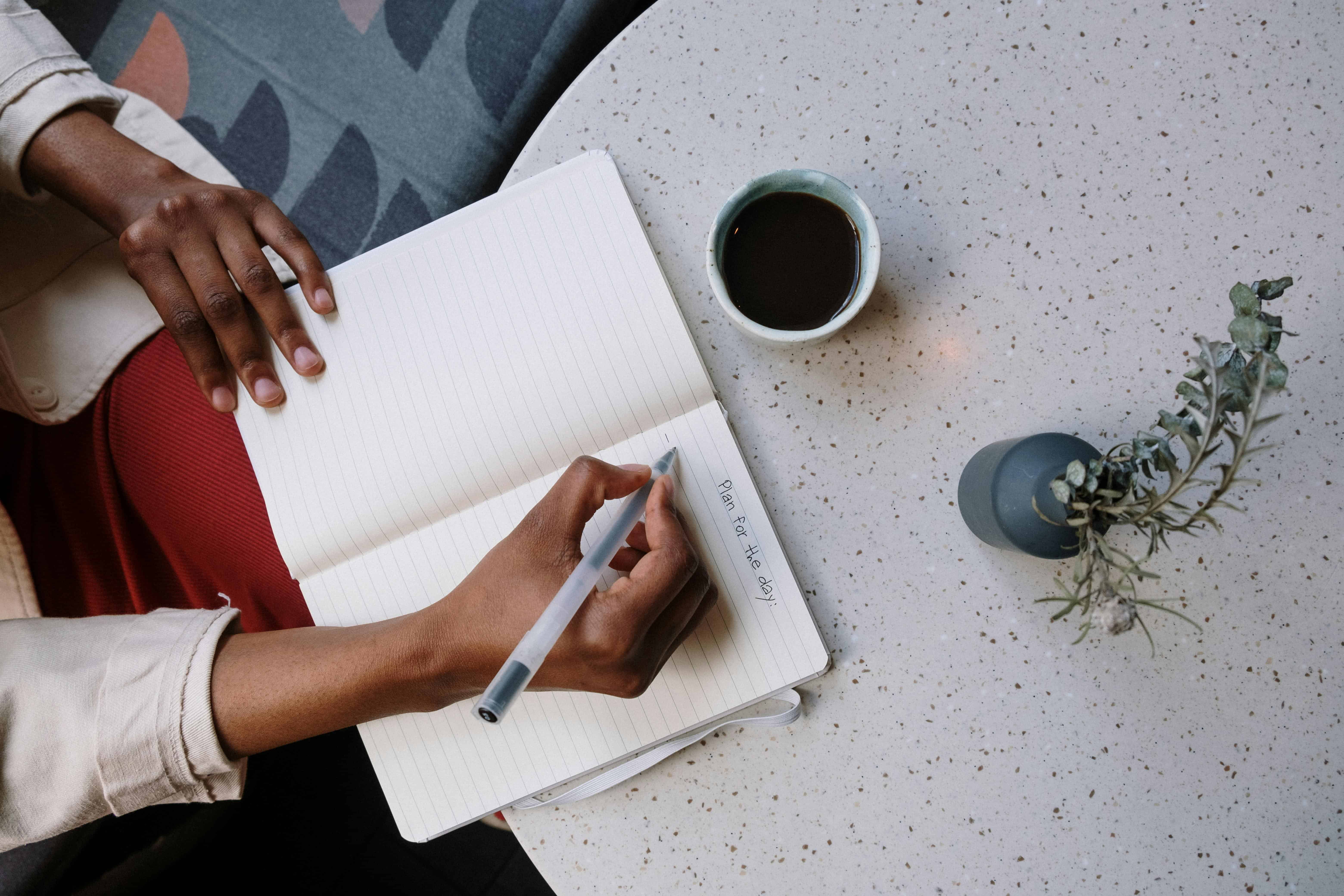 Person writing a daily plan in a notebook beside a cup of coffee, symbolizing habit-building and intentional routines