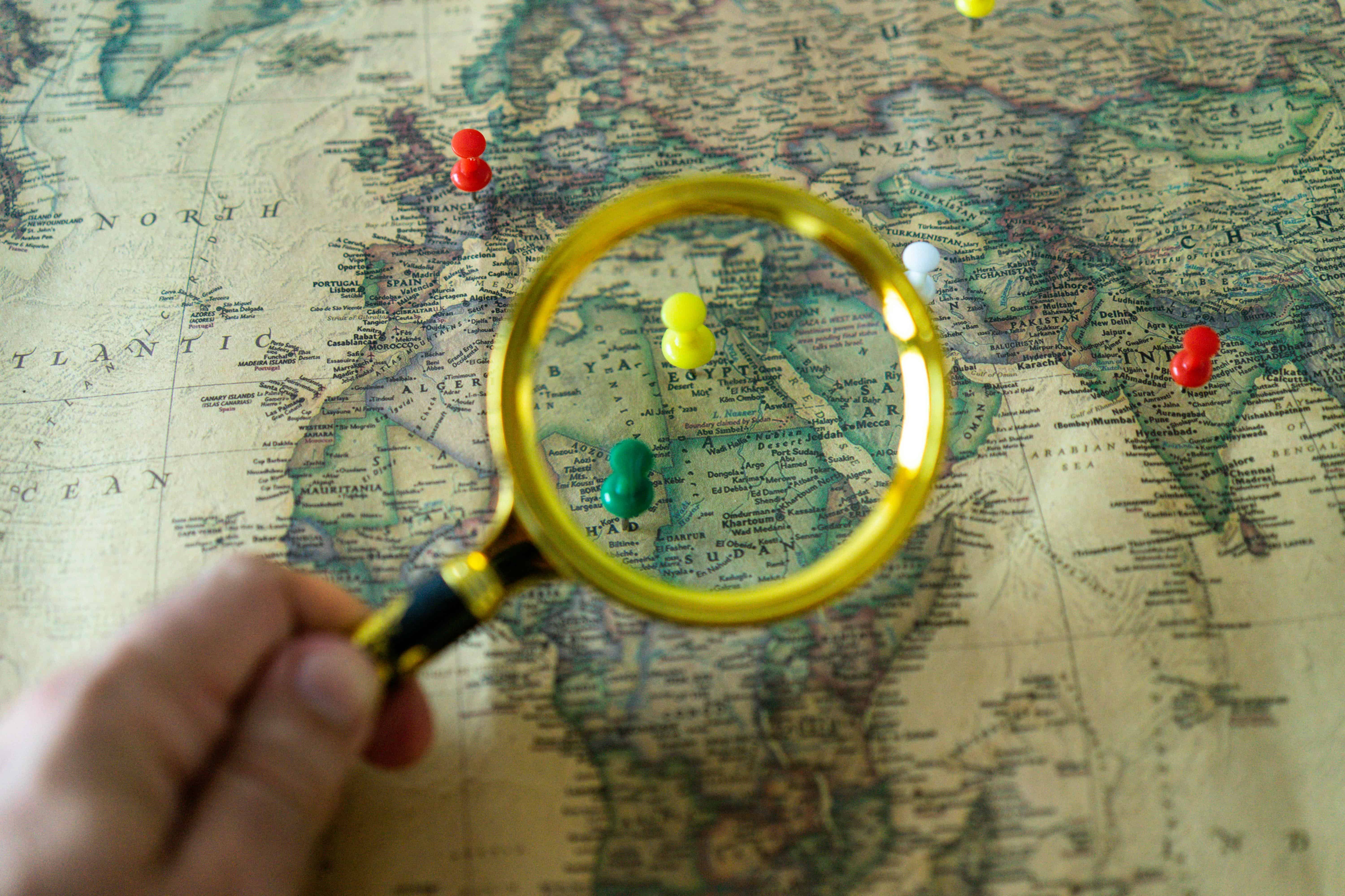 Hand holding a magnifying glass over a world map with colored pins marking destinations