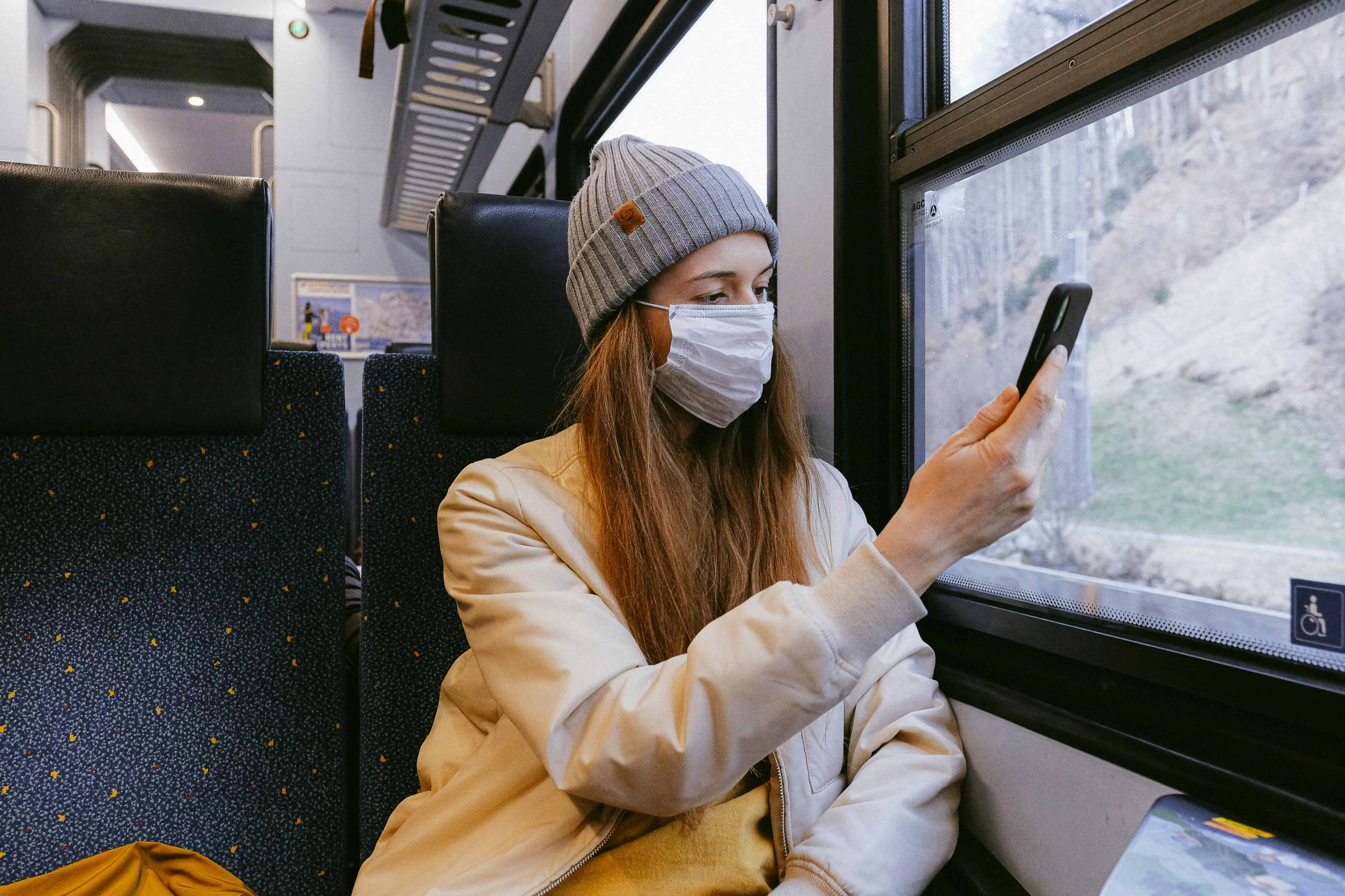 Woman wearing a face mask and beanie using her phone while traveling alone on a train