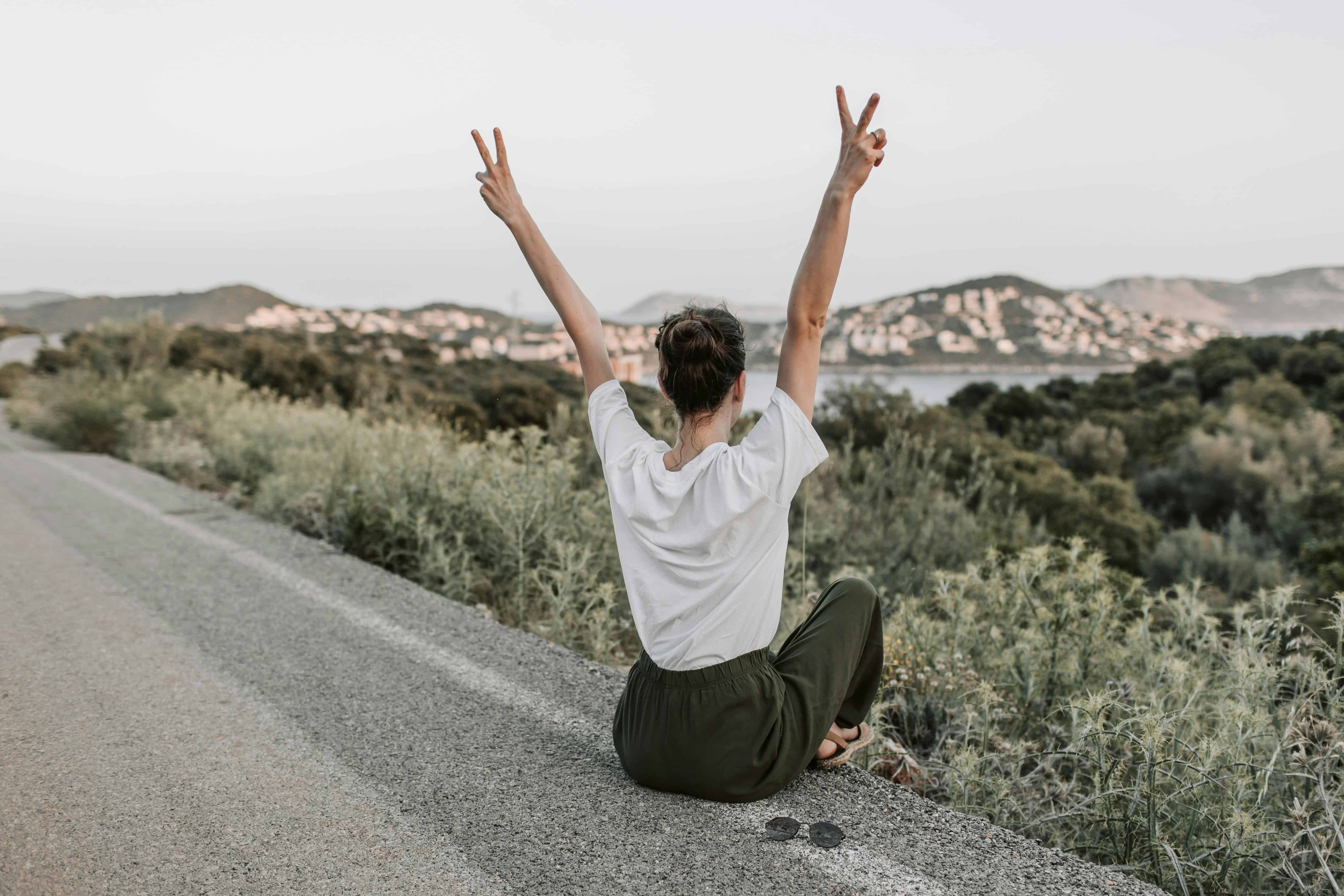 Woman sitting by the roadside with arms raised in peace signs, celebrating independence during solo travel.