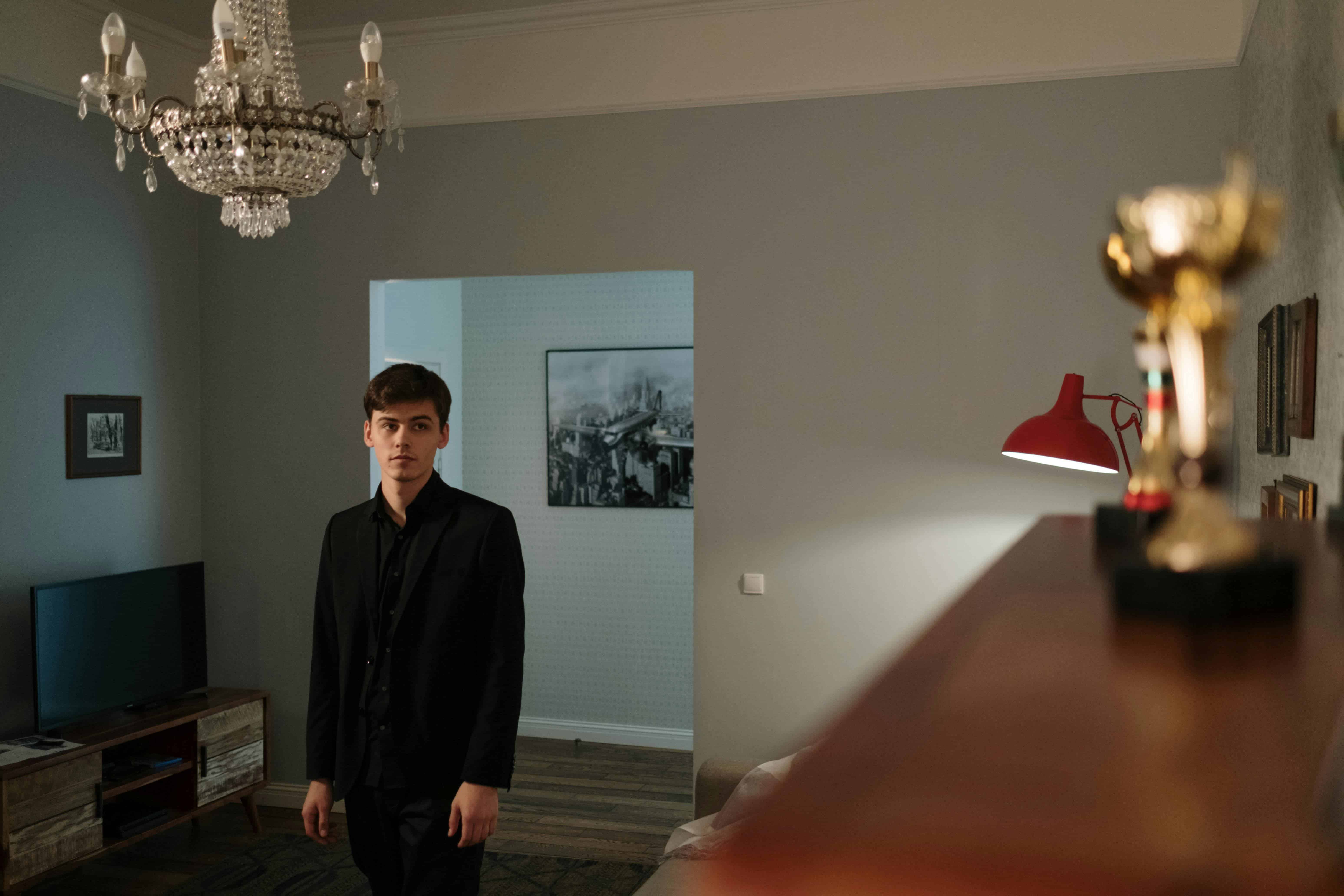 A man in a dark suit standing alone in a quiet room, facing a trophy in the foreground, symbolizing feeling empty after achieving success